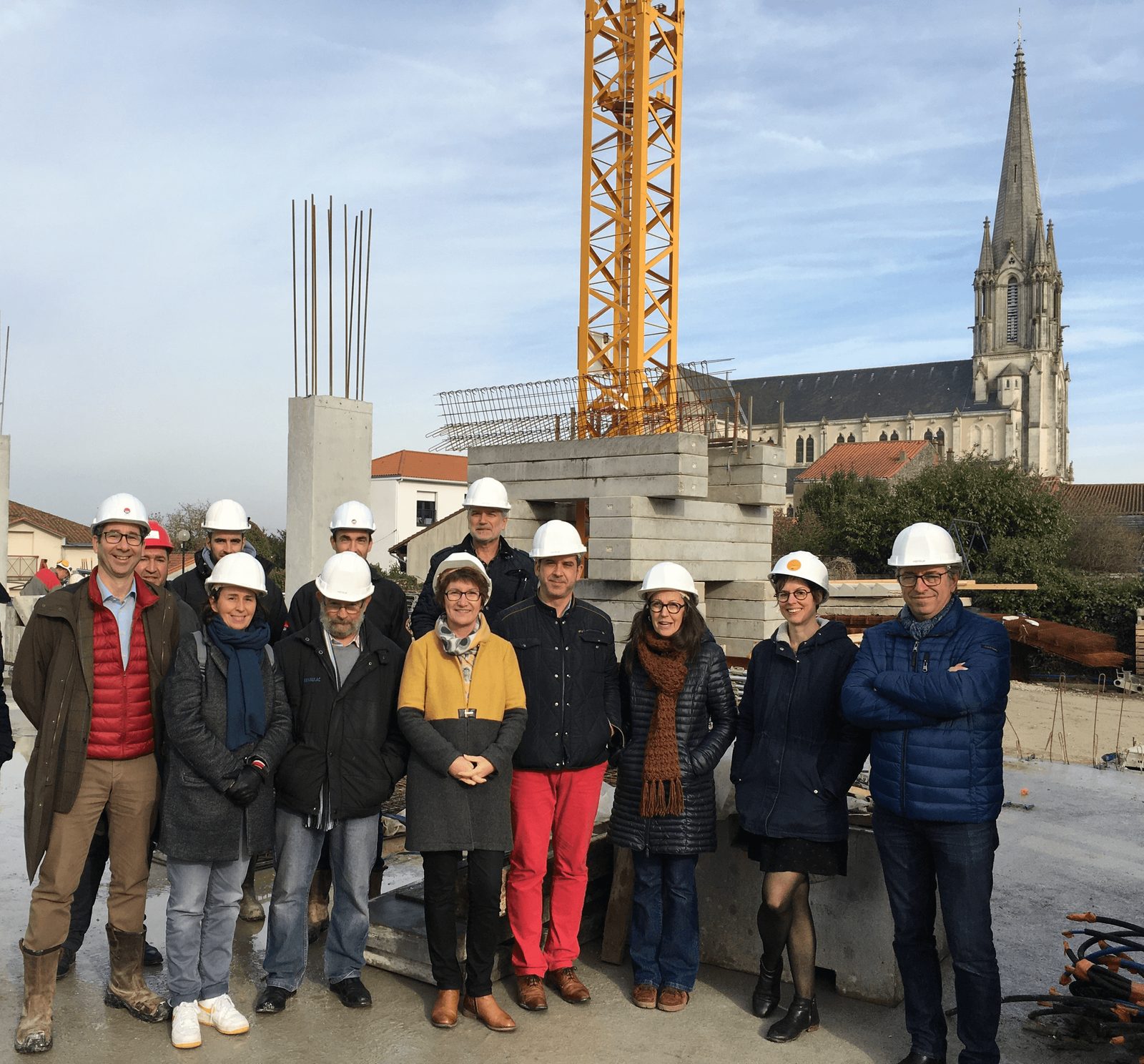 Visite de chantier avec les parties prenantes d'une opération, FedeA Visite de chantier en tenue de sécurité : intervenants réunis devant les poteaux en béton et la grue, église en arrière-plan
